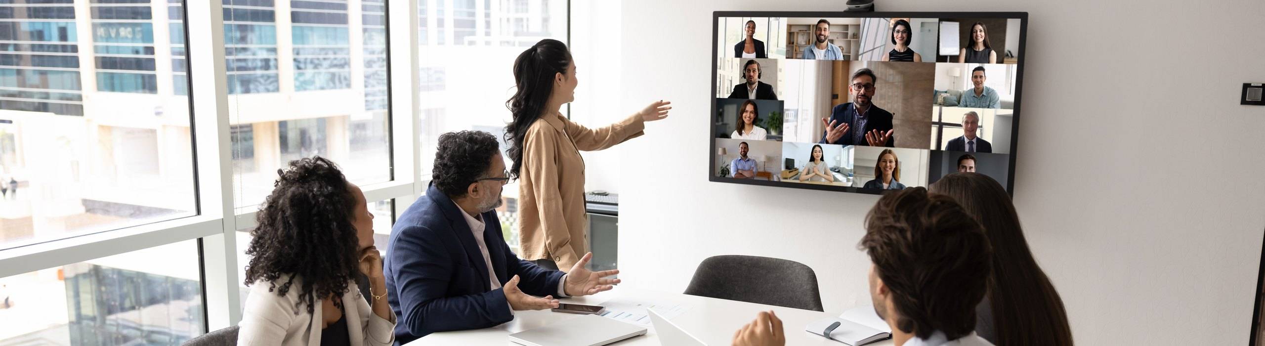 Colleagues in a meeting room presenting to remote participants shown on a wall-mounted video conferencing display