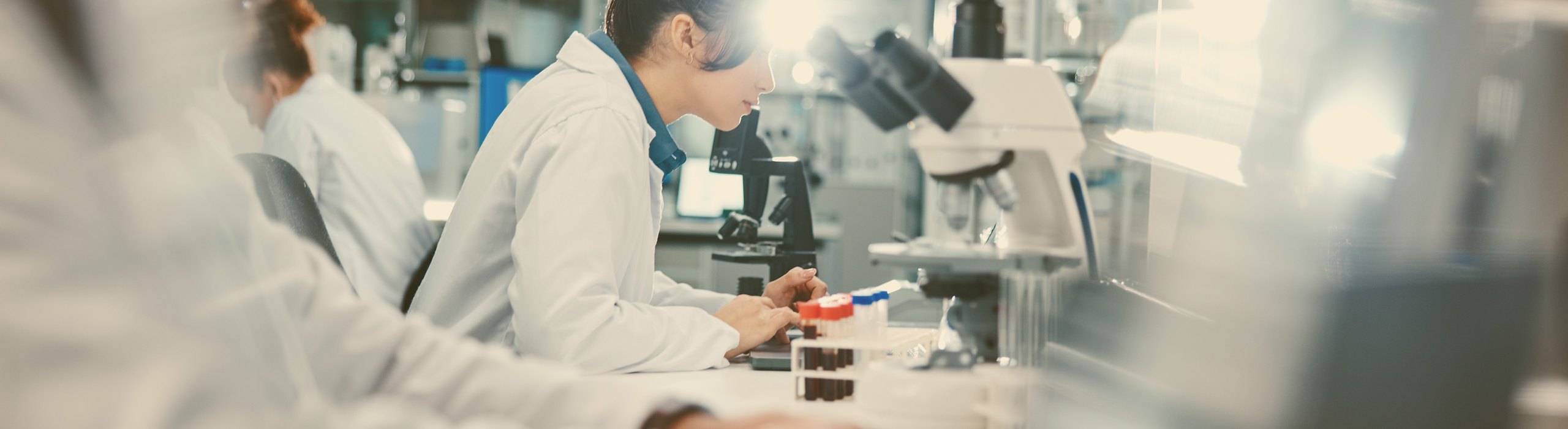 Researchers in white coats working at microscopes and sample racks along a laboratory bench.