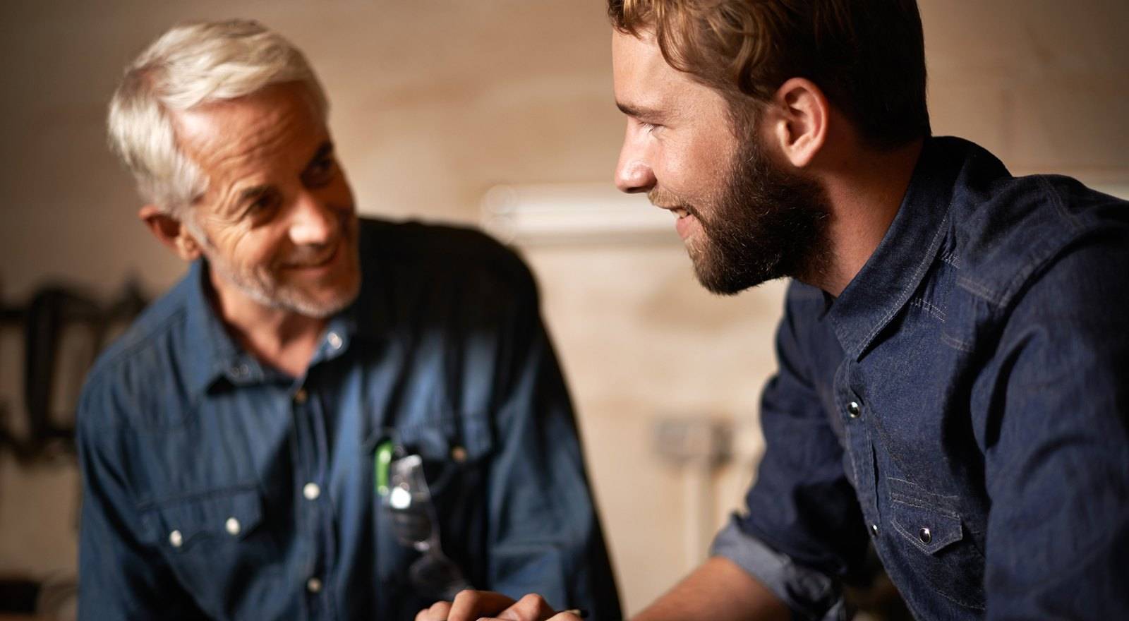Older business owner and younger man in denim shirts smiling and talking in a workshop, representing succession planning in a family business