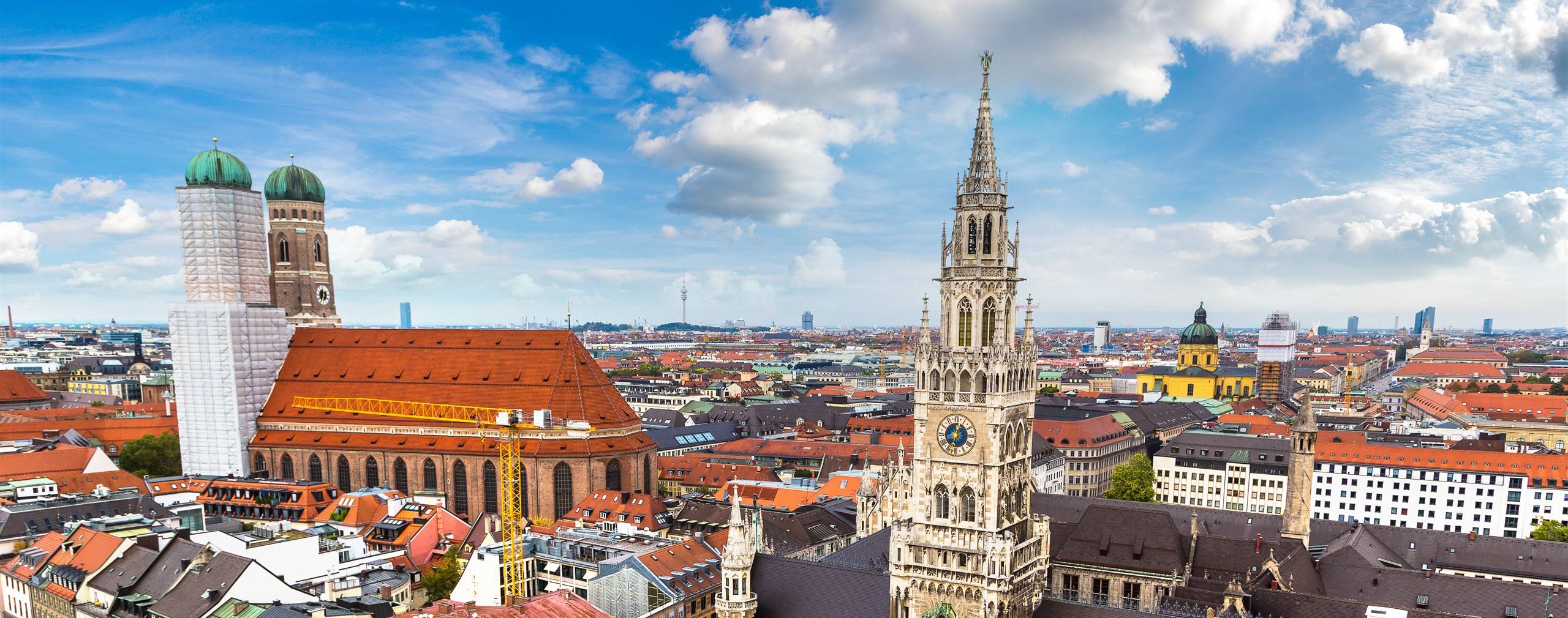 Aerial view across Munich's Marienplatz, the New Town Hall clock tower and the twin domes of the Frauenkirche rising above the city's red rooftops.