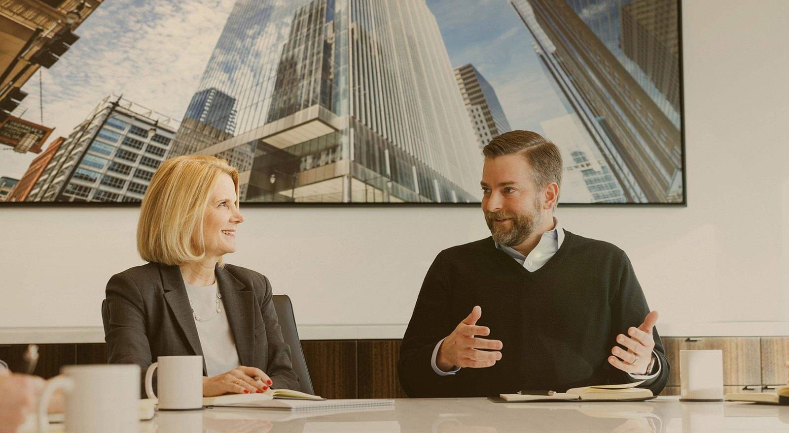 Two advisors, one in a charcoal suit and one in a dark sweater, speak at a conference table with notebooks and coffee mugs.