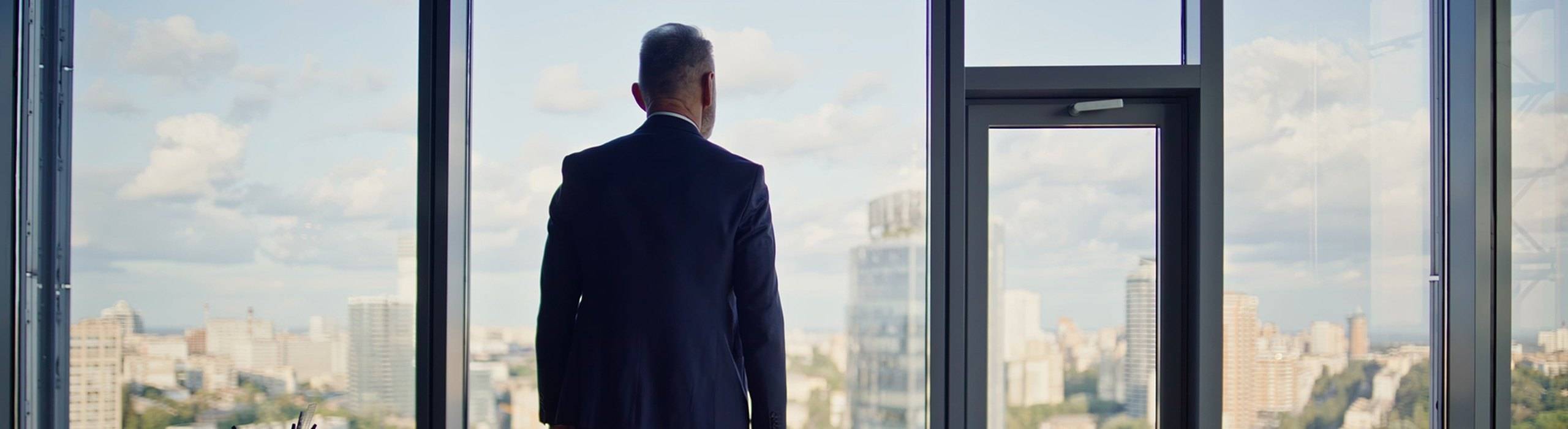 Business owner in a dark suit standing at a floor-to-ceiling office window overlooking the city, thinking about when to sell his business